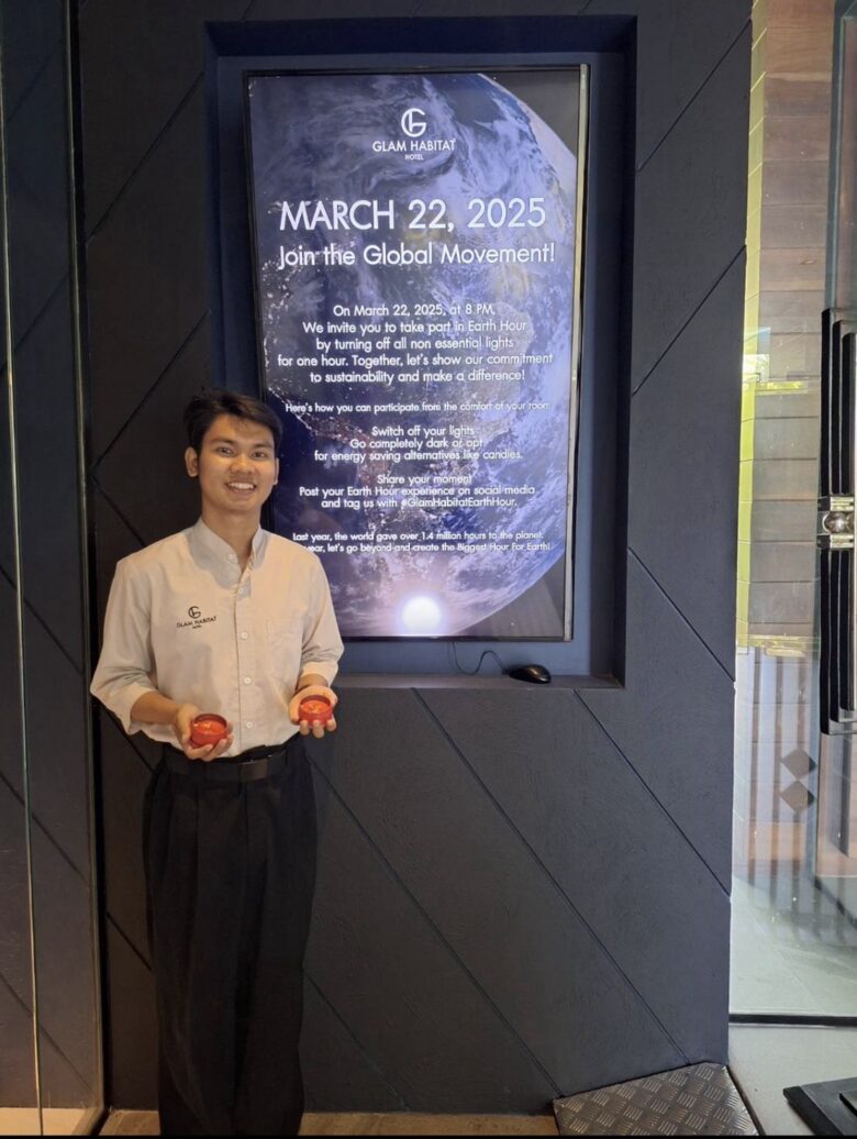 A Glam Habitat Hotel team member holding candles beside the Earth Hour 2025 invitation display at the hotel entrance