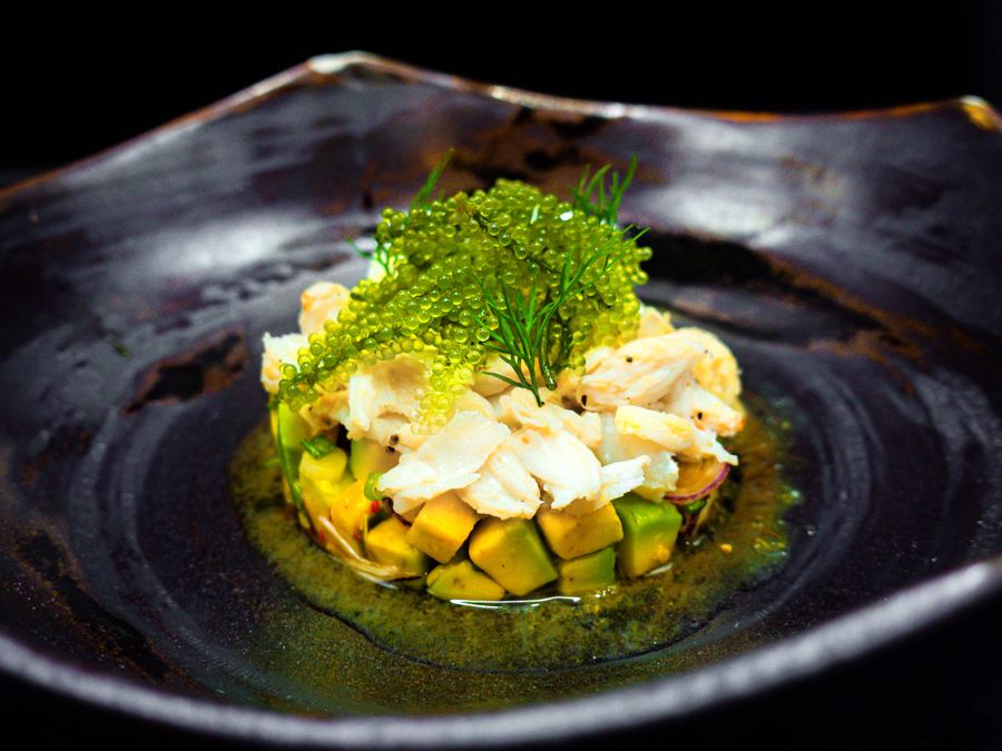 Seafood tartare with avocado and mango, topped with sea grapes, in a dark ceramic bowl